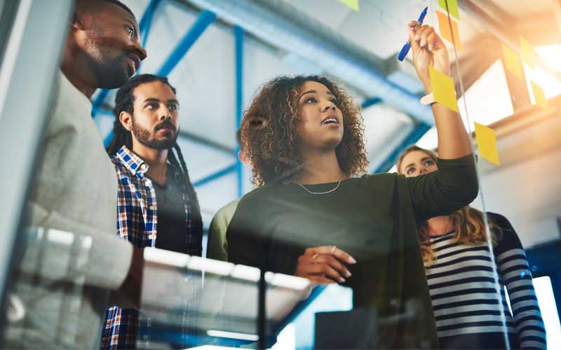 Un groupe de collègues aux profils variés collabore autour d'une paroi vitrée recouverte de post-it. Une femme écrit activement sur un post-it, sous le regard attentif des autres.