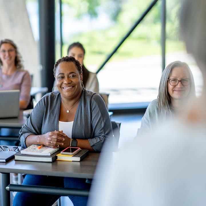 Un groupe de femmes d'horizons divers est assis à des tables, comme dans une salle de classe ou lors d'une réunion. La femme au premier plan arbore un large sourire et regarde le spectateur.