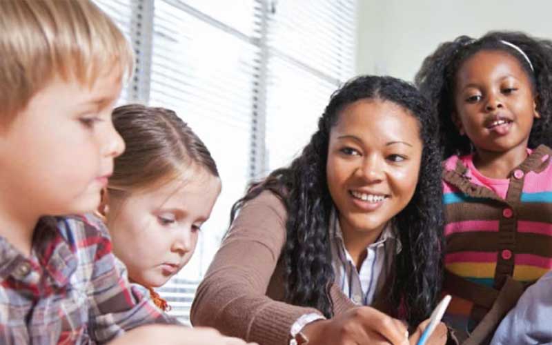 Une enseignante interagit avec trois jeunes élèves dans une salle de classe. Elle sourit et semble participer à une activité avec les enfants.