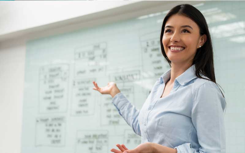 A smiling woman in a light blue shirt gestures towards a whiteboard with a flowchart on it. She appears to be presenting or teaching.