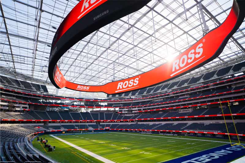 Intérieur d’un grand stade de football américain moderne, vide, avec un immense écran incurvé affichant « ROSS », des tribunes bleues à plusieurs niveaux, un terrain marqué et lumineux, et une structure de plafond laissant entrer la lumière naturelle.