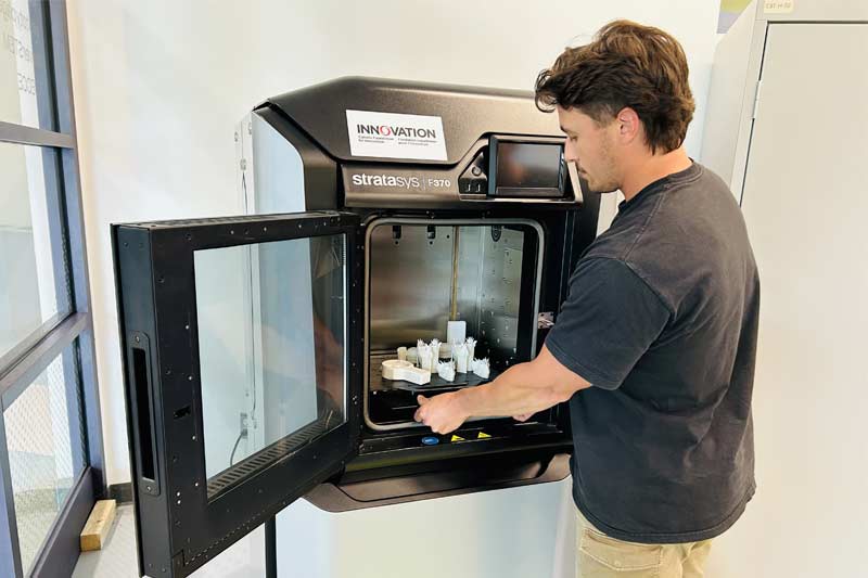 Homme manipulant une imprimante 3D industrielle Stratasys F370, retirant ou inspectant des objets imprimés dans un environnement moderne et bien éclairé.