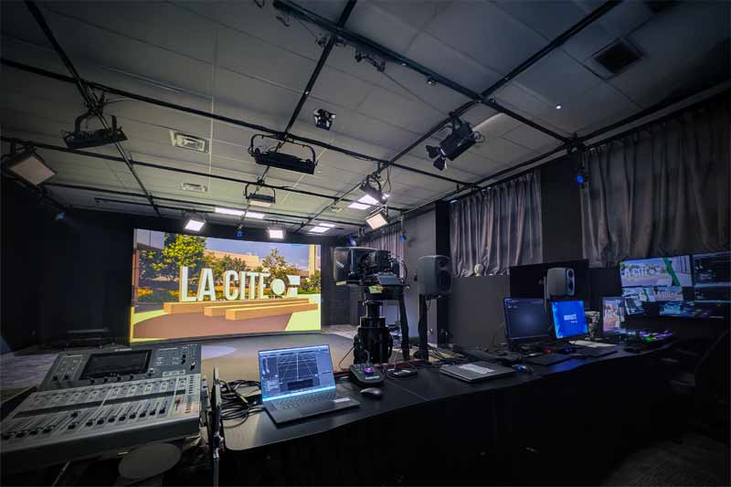 Intérieur d’un studio de télévision professionnel moderne, avec un grand écran central affichant « LA CITE », un bureau de contrôle avec équipements et caméras, éclairage studio sophistiqué et murs sombres contrastant avec le décor lumineux.