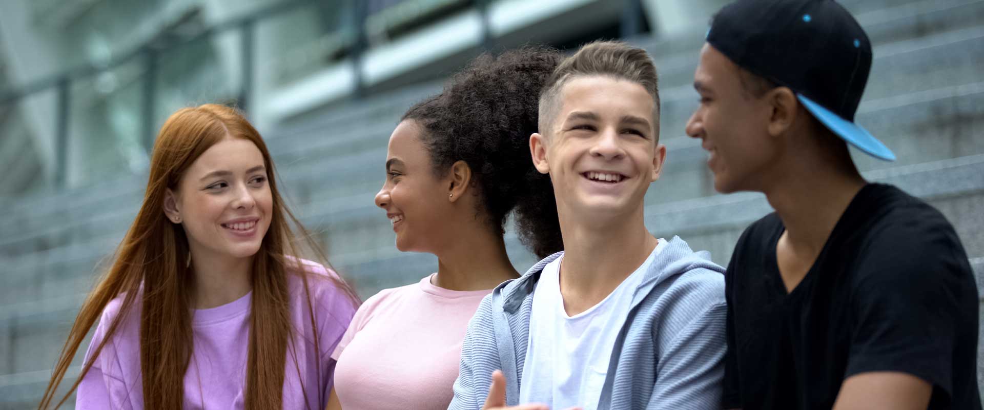 Four teenagers from different backgrounds are sitting on steps and smiling. They seem to enjoy each other's company.