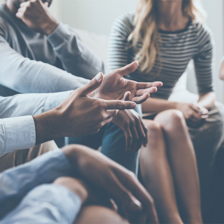 Gros plan sur plusieurs personnes assises en pleine conversation, l'accent étant mis sur leurs mains et leurs gestes. L'image suggère une discussion ou une réunion.