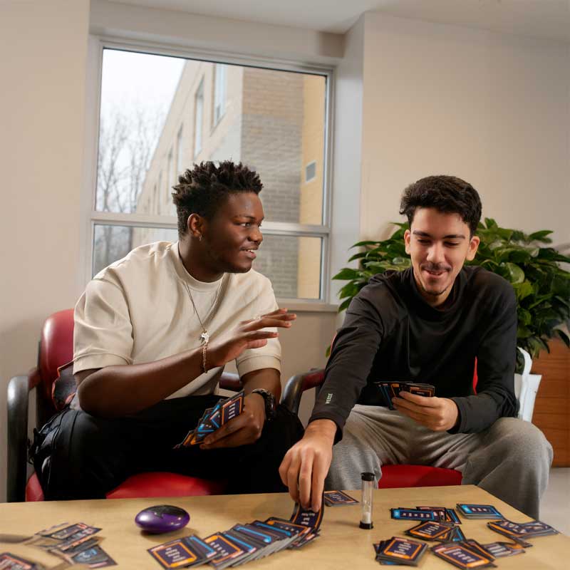 L'image montre deux jeunes hommes assis autour d'une table, en train de jouer à un jeu de cartes. On dirait qu'ils s'amusent et qu'ils sont concentrés sur le jeu. L'arrière-plan est flou, mais on peut voir qu'ils sont probablement à l'intérieur, peut-être dans une salle de jeu ou un salon.